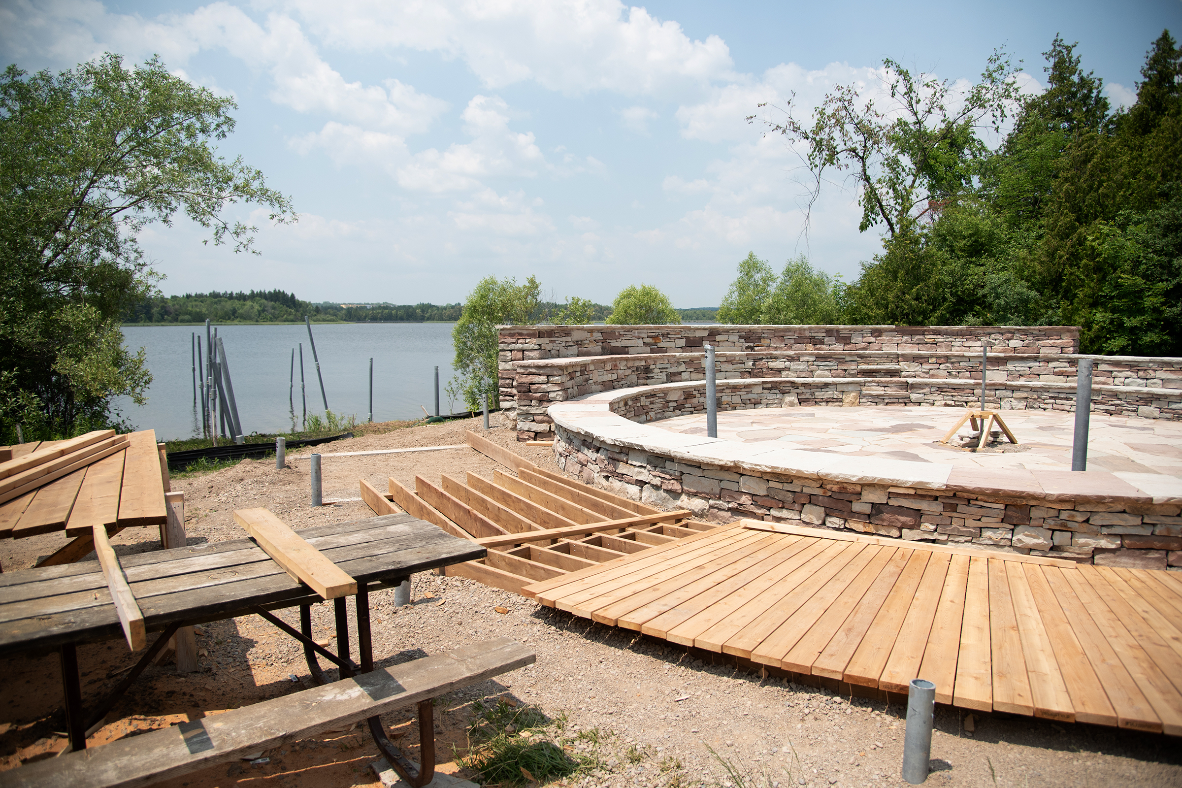 Construction of the Crane Gathering Space. Alt text: A round amphitheatre made of stone with surrounded by a wooden boardwalk under construction with a lake in the background
